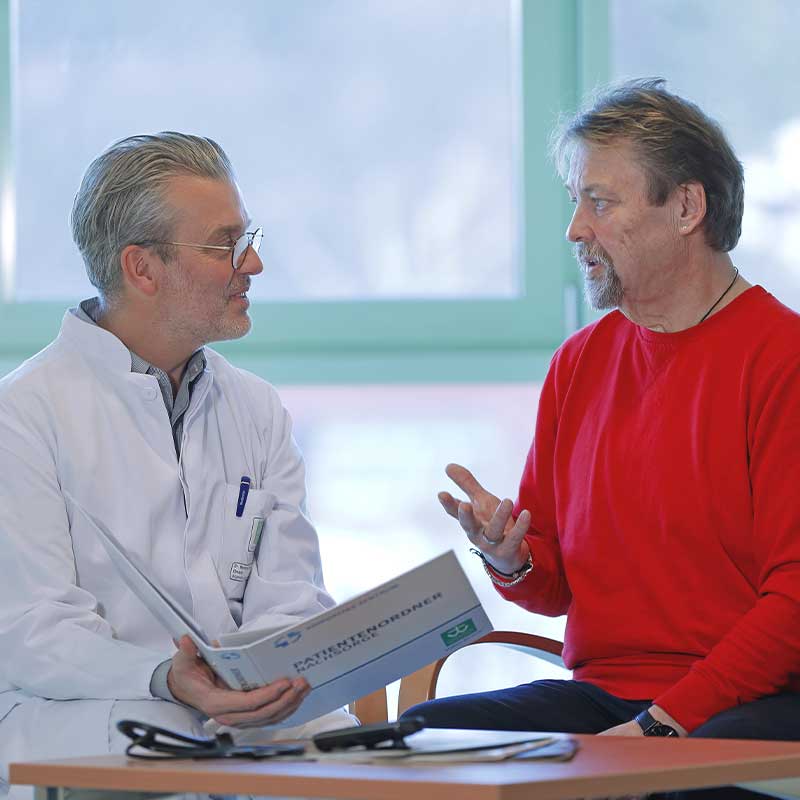 Ein Arzt und ein Patient im Gespräch. Der Arzt hält einen Patientenordner.