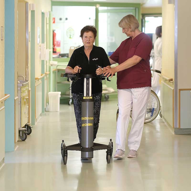 Eine Frau in einem Korridor benutzt ein Steh- und Gehhilfegerät, begleitet von einer Gesundheitsfachkraft.