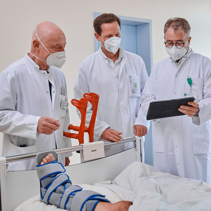 Ärzte in weißen Kitteln und Masken stehen an einem Krankenbett, prüfen ein Tablet; Bein des Patienten ist bandagiert.