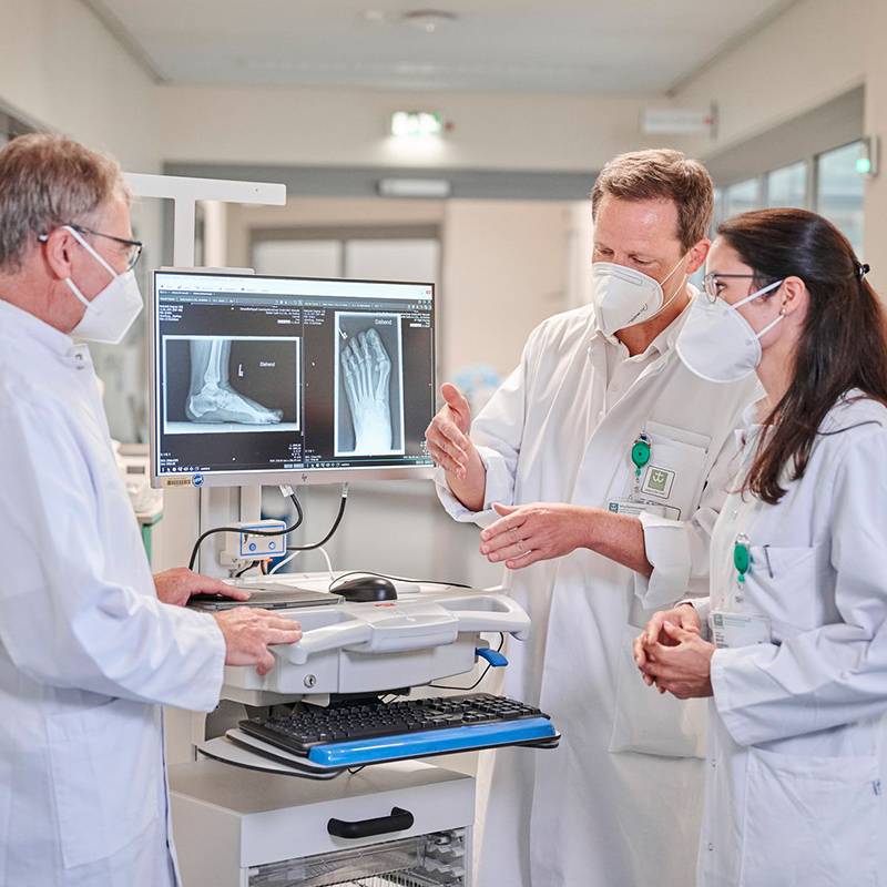 Drei Ärzte im Gespräch, betrachten Röntgenbilder auf einem Bildschirm. Alle tragen weiße Kittel und Masken.