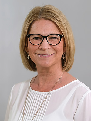 Es ist ein Porträt einer lächelnden Person mit Brille und weißem Oberteil vor grauem Hintergrund.