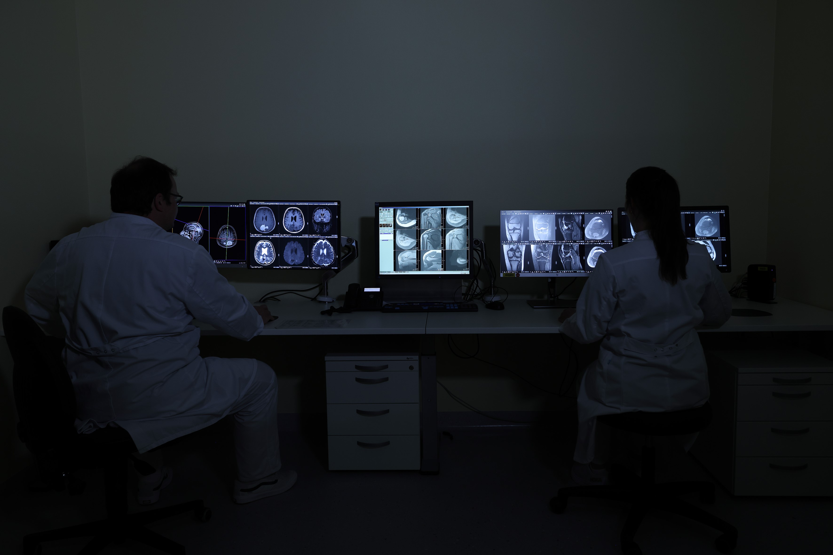 Zwei Personen in weißen Kitteln betrachten medizinische Scans auf mehreren Monitoren in einem dunklen Raum.